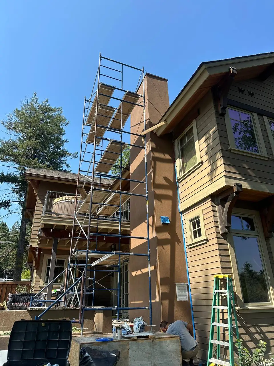 Applying a warm brown custom stucco finish to a multi-story exterior chimney using scaffolding, contrasting with wood siding